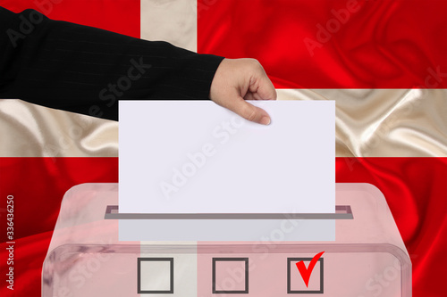 female voter lowers the ballot in a transparent ballot box against the background of the national flag of Denmark, concept of state elections, referendum