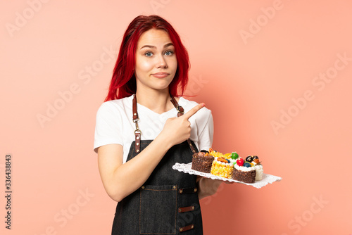 Pastry chef holding a muffins isolated on pink background pointing to the laterals having doubts