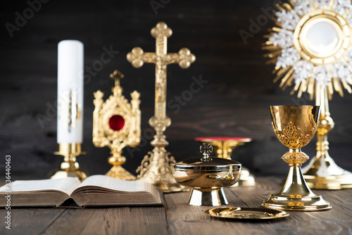 Catholic religion concept. Catholic symbols composition. The Cross, monstrance, Holy Bible and golden chalice on wooden altar and gray background. 