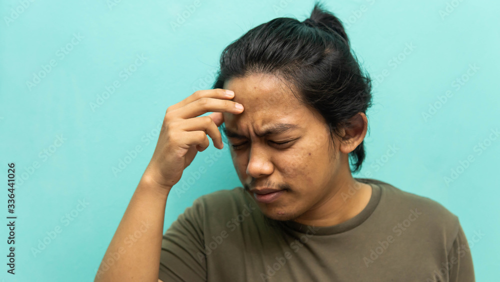 A portrait of a Malay man holding his head, having a headache with painful face reaction on isolated blue background. A portrait of a Malay man holding his head, having a headache with painful face reaction on isolated blue background.