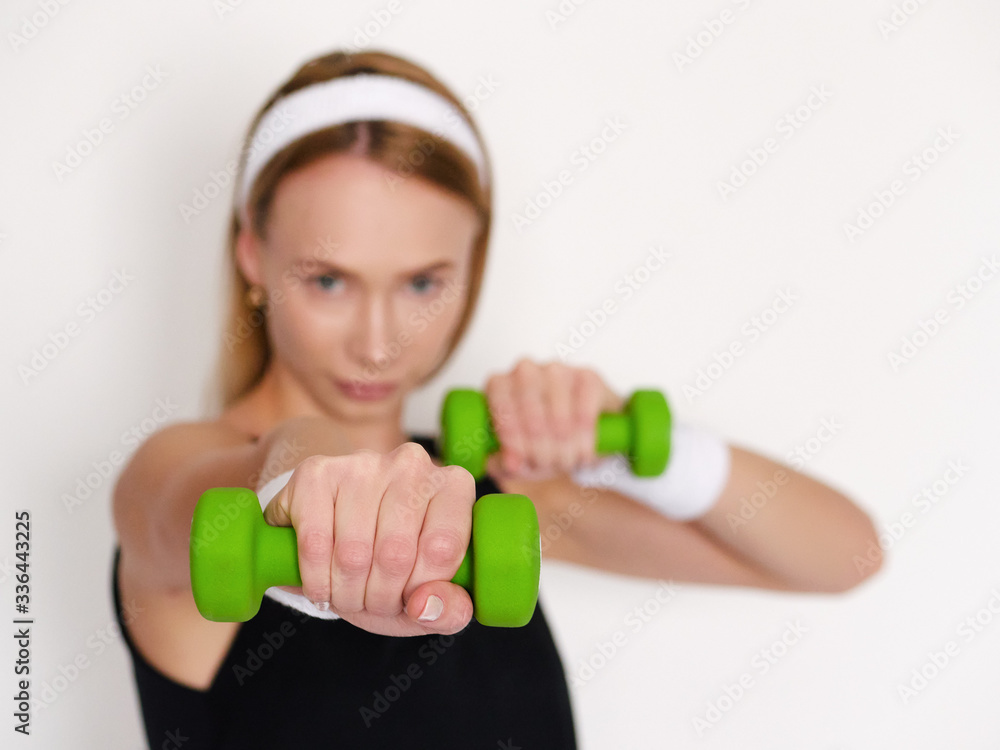 Beautiful young sporty woman in black outfit doing exercises with green dumbbells to stay healthy and active on self isolation in home quarantine posing against white wall focus on dumbbell Beautiful young sporty woman in black outfit doing exercises with green dumbbells to stay healthy and active on self isolation in home quarantine posing against white wall focus on dumbbell