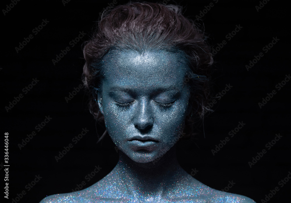 girl with a blue face in sparkles close-up against a black wall 