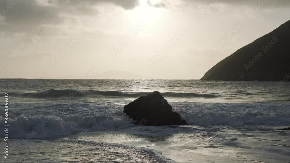 One strong boulder lying on a remote beach, waves crashing into it causing foamy waves and spray in glow coming from sun behind clouds. Achill Island. One strong boulder lying on a remote beach, waves crashing into it causing foamy waves and spray in glow coming from sun behind clouds. Achill Island.