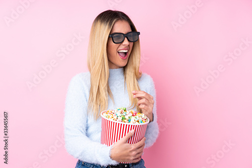 Young Uruguayan woman over isolated pink background with 3d glasses and holding a big bucket of popcorns while looking side