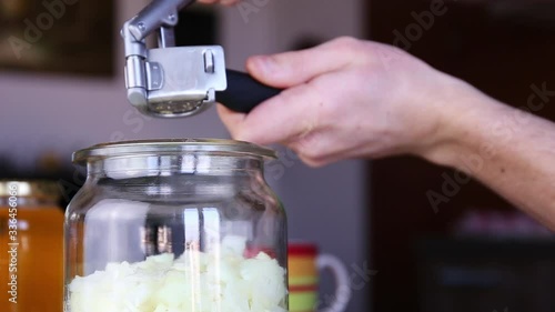 Male hands use garlic presser to press garlic cloves to a glass jar with pieces of onion 