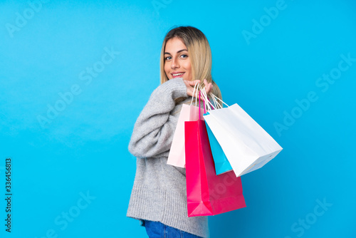 Fotografi Young Uruguayan woman over isolated blue background holding shopping bags and sm