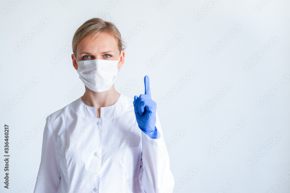Attention. Blonde female doctor surgeon in white uniform, protective blue gloves and medical mask showing finger up over grey Attention. Blonde female doctor surgeon in white uniform, protective blue gloves and medical mask showing finger up over grey