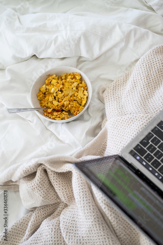 Kempton Park, Gauteng / South Africa - 04/07/2020: Man sits in bed working at home on his apple mac while eating breakfast in bed, scrambled eggs in a bowl, white bedsheets photo