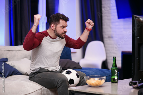 Good-looking modern positive 30-aged guy with well-groomed beard savoring chipps during emotional review of favourite football team's game on tv at home photo