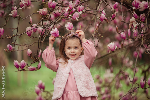 Portrait of a cute cheerful girl in a pink dress wearing a fur vest who smiles on a walk in spring in a park under a flowering pink magnolia tree.