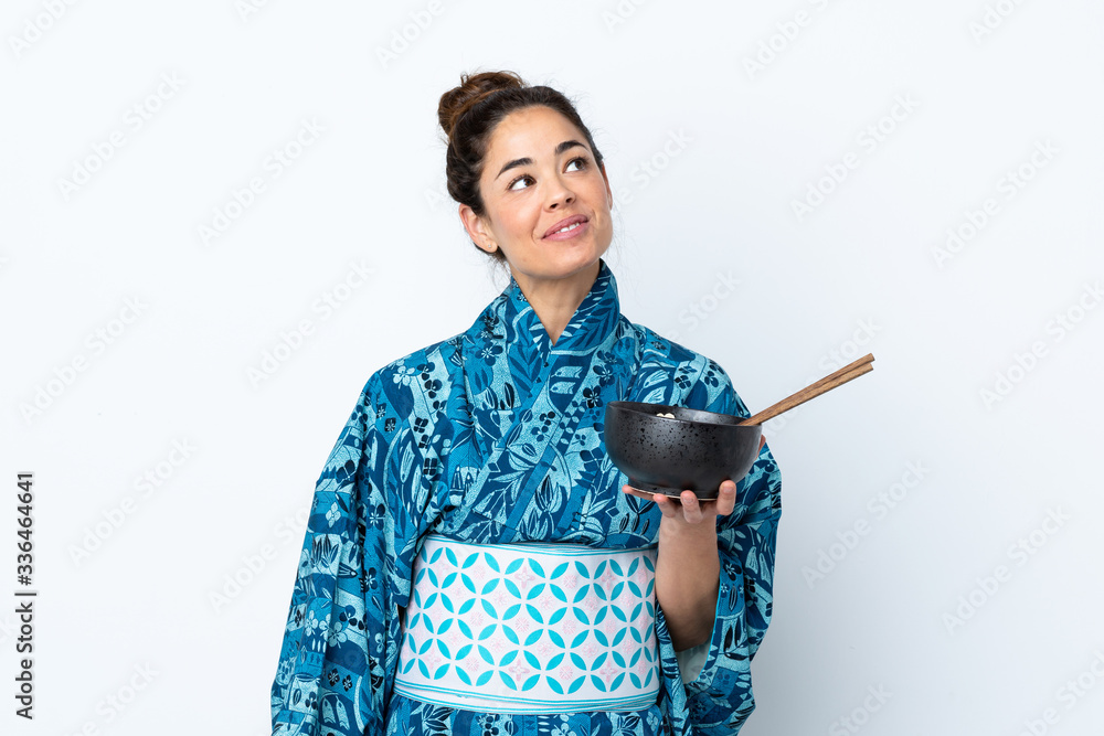 Woman wearing kimono over isolated white background looking up while smiling while holding a bowl of noodles with chopsticks Woman wearing kimono over isolated white background looking up while smiling while holding a bowl of noodles with chopsticks