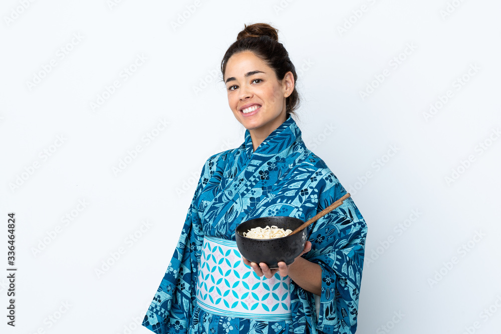 Woman wearing kimono over isolated white background smiling a lot while holding a bowl of noodles with chopsticks Woman wearing kimono over isolated white background smiling a lot while holding a bowl of noodles with chopsticks