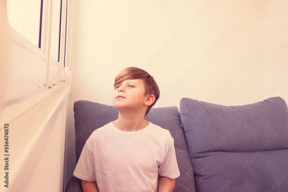 Child boy looking outside sitting on grey stylish sofa near window in modern interior and missing friends or dreaming about future, staying home Child boy looking outside sitting on grey stylish sofa near window in modern interior and missing friends or dreaming about future, staying home