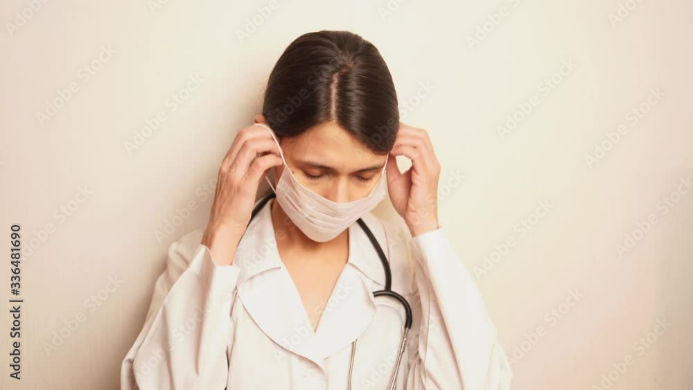 a pretty female doctor looks seriously at the viewer, takes a deep breath and puts on a white disposable protective mask and disposable gloves, preparing to treat patients a pretty female doctor looks seriously at the viewer, takes a deep breath and puts on a white disposable protective mask and disposable gloves, preparing to treat patients