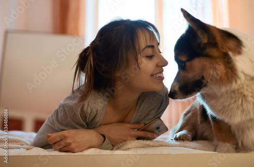 Close-up portrait of pleased woman lying on bed at home with her loved dog Welsh Corgi, enjoys morning weekend together. Concept stay at home, friendship with pet, meet morning in bed.
