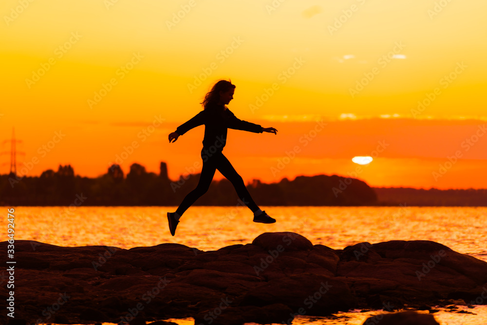 Young slim woman jumping on rock on sunset background. Nature and beauty concept. Orange sundown. Girl silhuette at sunset.