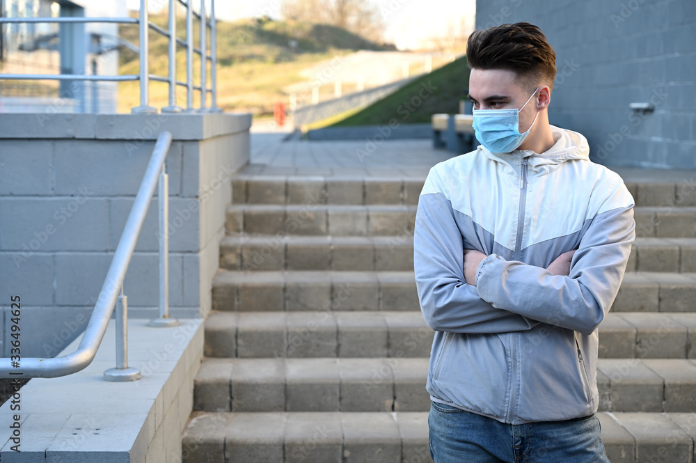 Fototapeta premium Photo Caucasian young man in a medical mask in a light jacket outdoors on a city background on a sunny day
