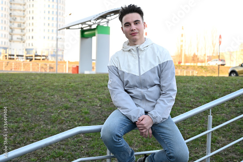 Portrait of a Caucasian young brunette man in a light jacket outdoors on a city-background. Model is sitting on the railing of the stairs
