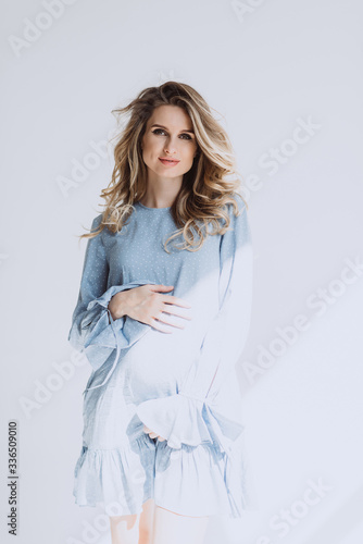 Beautiful pregnant woman with curls and make-up in a light blue dress on a white background in the rays of the sun hugs her belly. Waiting for a child. Soft selective focus. © Tasha Sinchuk