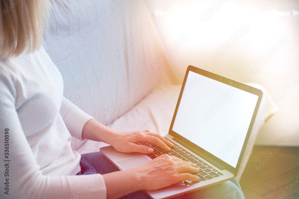 Naklejka premium Portrait of blonde hair woman using her blank screen laptop while sitting on the sofa at home in the sunset light. Blonde female sit indoors at home working on computer with blank screen. Home office
