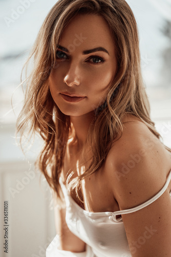 Close up portrait of an elegant and sexy female model with brown curly hair wearing white lace lingerie and shirt while sitting in a bright apartment, looking thoughtful