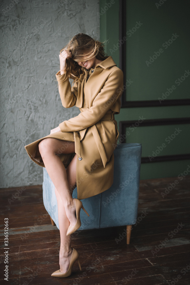 Beautiful sexy woman with curls and chubby lips and long legs in a beige coat in a loft style studio on a blue armchair. Beauty and fashion. Soft selective focus.