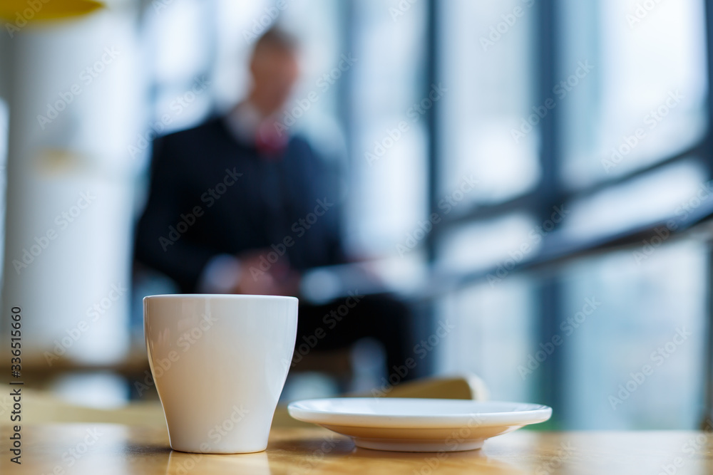 A white cup and saucer with hot aromatic coffee stands on a wooden table against the background of a sitting businessman