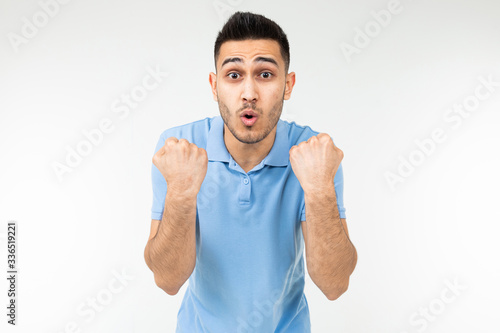 handsome Caucasian in a blue T-shirt rejoices victory holding his fists on a white studio background