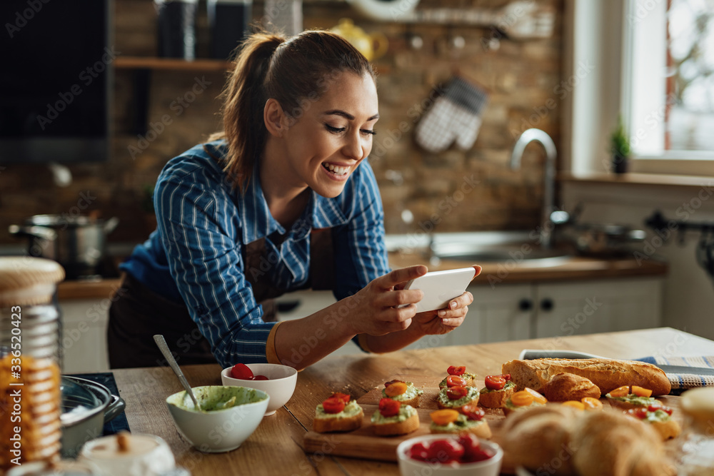 Happy woman using smart phone and photographing food she has prepared in the kitchen.