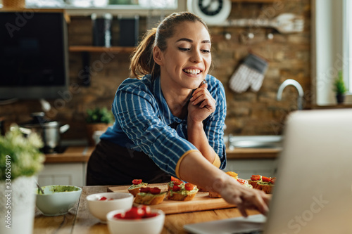 Happy woman surfing the net on laptop while preparing bruschetta in the kitchen.
