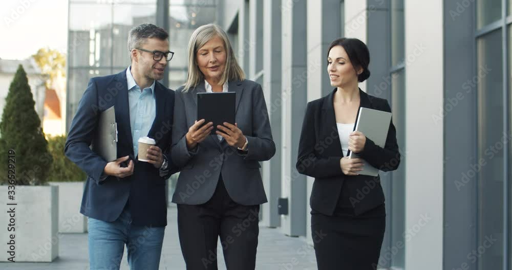 Caucasian gray-haired woman boss walking the street with male and female workers and showing them project on tablet device screen. Man and woman stepping outdoor with head of company. Caucasian gray-haired woman boss walking the street with male and female workers and showing them project on tablet device screen. Man and woman stepping outdoor with head of company.