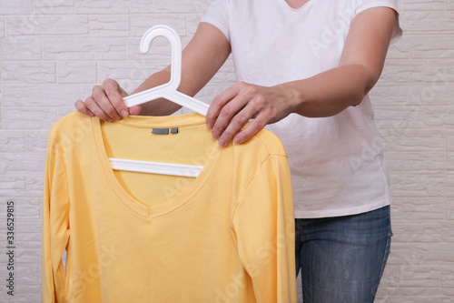 Caucasian woman choosing clothes, she is holding a hanger with yellow longsleeve, shopping, fitting and buying clothes during sale and discount concept, cheap second hand clothes for online selling