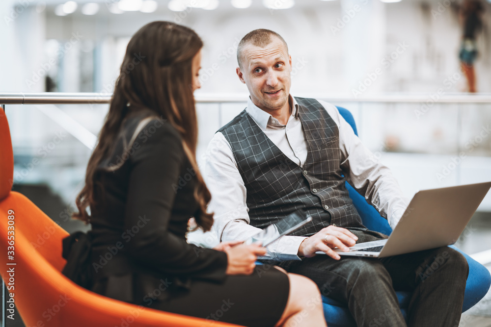 A business talk in an open space area of a contemporary office: a mature man entrepreneur with a laptop on his knees is lively setting tasks to his female partner next to him, multi-colored armchairs A business talk in an open space area of a contemporary office: a mature man entrepreneur with a laptop on his knees is lively setting tasks to his female partner next to him, multi-colored armchairs