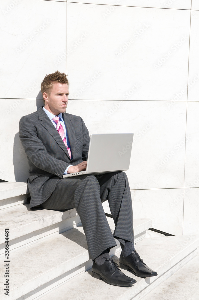 Serious businessman working remotely sitting outdoors on sunny white steps using his laptop computer Serious businessman working remotely sitting outdoors on sunny white steps using his laptop computer