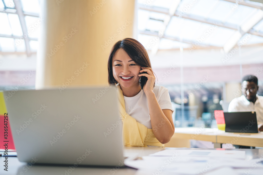 Asian woman smiling during cellphone conversation in modern office interior, happy Japanese female professional discussing ideas for project using mobile phone for calling and laptop for info browsing Asian woman smiling during cellphone conversation in modern office interior, happy Japanese female professional discussing ideas for project using mobile phone for calling and laptop for info browsing