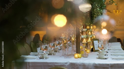 Square table served with wine glasses, cutlery and glassware, decorated with stylish candlesticks, and green branches with leaves