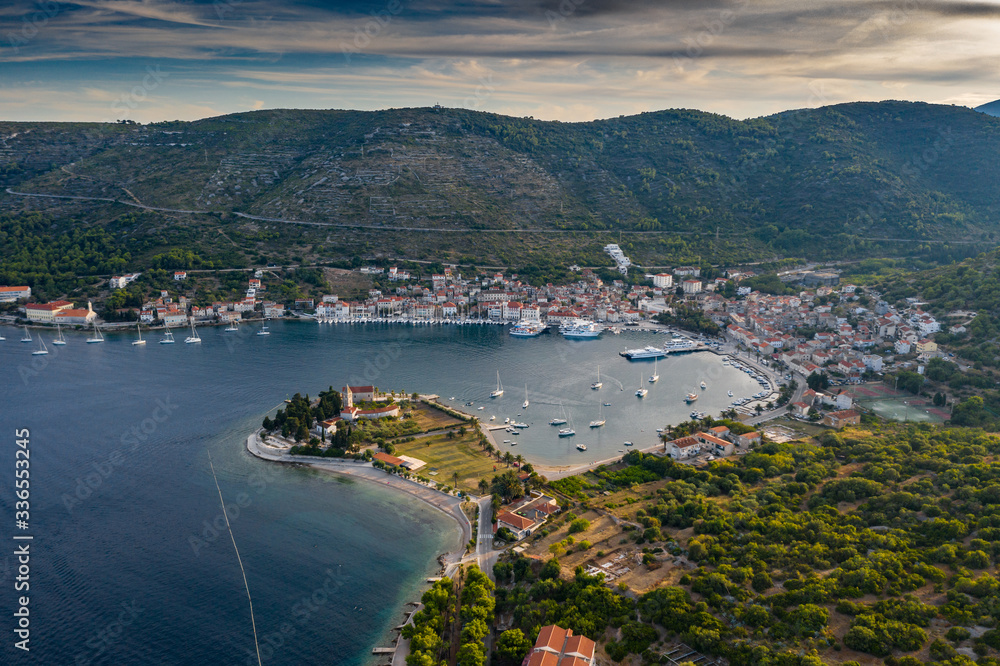 Aerial view of marina Vis at sunset, Croatia, a lot of chaotically standing boats in a bay, roofs of orange color, sunshine, hills with green trees, ferry station