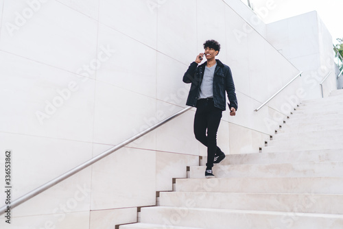 Positive man in fashionable outfit smiling during smartphone cell call with roaming internet connection, successful male millennial in trendy apparel communicating via digital mobile phone