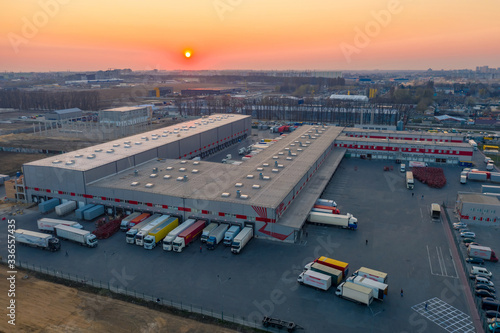 Aerial view of the logistics park with warehouse, loading hub and many semi trucks with cargo trailers standing at the ramps for load/unload goods at sunset