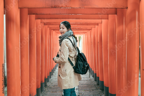 side view of beautiful asian chinese woman walking through famous lines of orange column Torii at tenmangu temple in osaka. girl backpacker holding camera turn face around standing under red gates