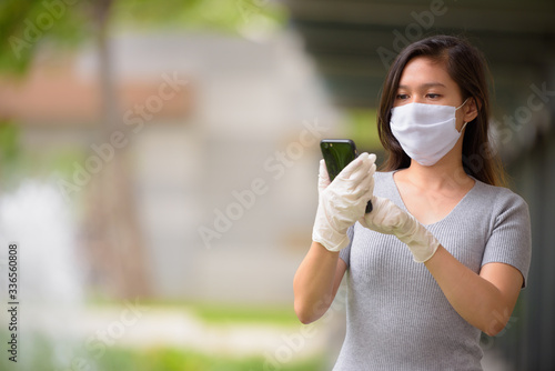 Young Asian woman wearing mask and gloves while using phone outdoors
