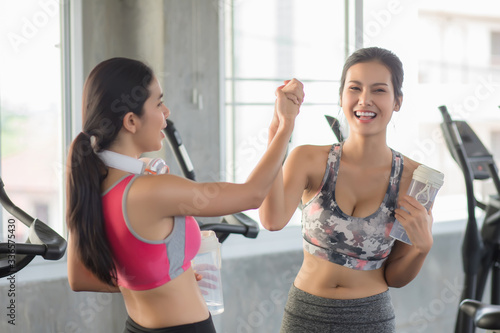 Two beautiful Asian women, wearing exercise clothes, holding a water bottle and holding hands in the gym filled with exercise equipment with bright smiles. In the concept of achieving fitness goals