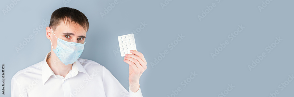 Banner.  A man in a medical mask holds pills on a gray background with emotions, dressed in a white shirt, uniform, on the right there is an empty space for the text. Coronavirus, Covid-19