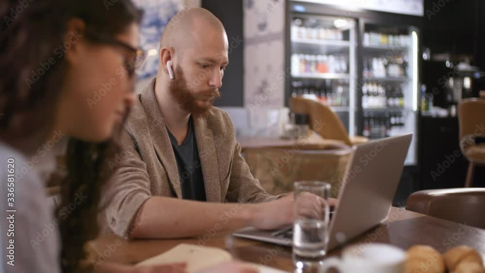 Caucasian businessman in wireless headphones typing on laptop at table in coffeeshop while female colleague taking notes Caucasian businessman in wireless headphones typing on laptop at table in coffeeshop while female colleague taking notes