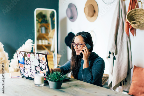 Young charming stylish woman in a home office sits at a desk, looks at a laptop and speaks while smiling on the phone. The concept of freelance and remote work. Stay safe and self-isolation. photo