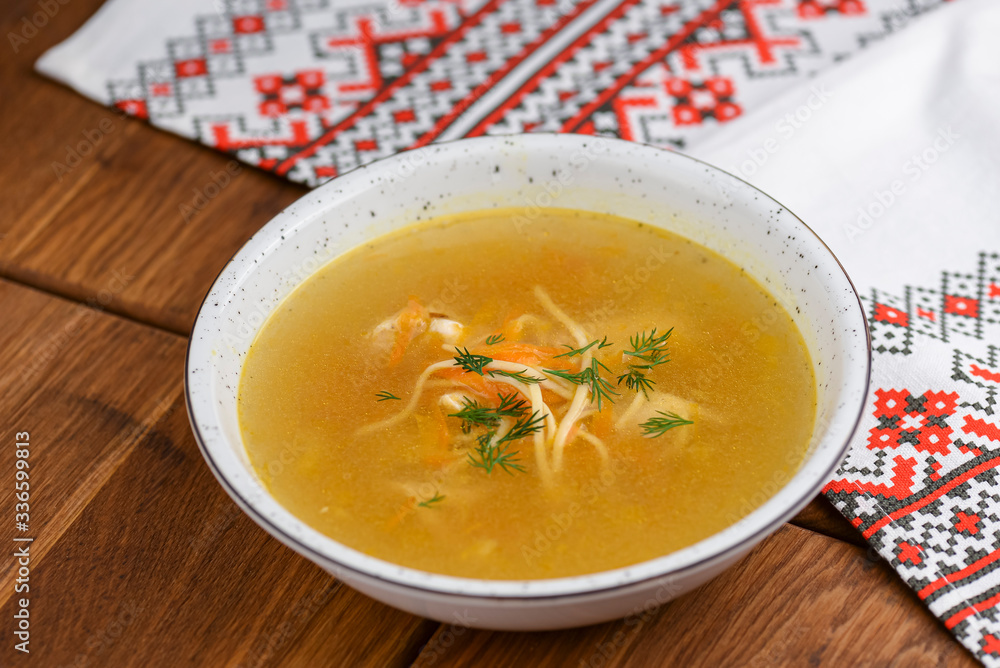 Chicken soup with noodles and vegetables served in white bowl over rustic wooden table with traditional tablecloth. Chicken soup with noodles and vegetables served in white bowl over rustic wooden table with traditional tablecloth.