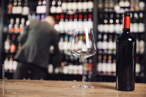 Bottle red wine and glass goblet on wooden table, in background sommelier chooses drink from restaurant shelf