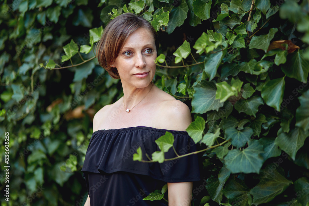 Attractive woman in a black dress with bare shoulders and a short haircut poses to the will of a bright green wall of plants. Attractive woman in a black dress with bare shoulders and a short haircut poses to the will of a bright green wall of plants.