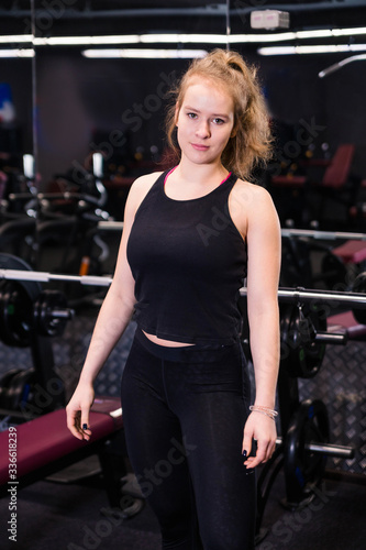 Front view of female bodybuilder posing near sports equipment, in gym in dark atmosphere, looking at camera.....