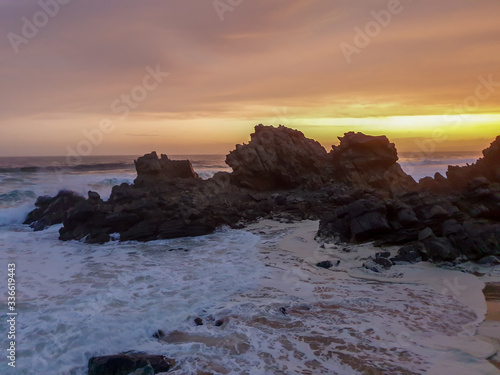 Sea splashing around jagged rocks on the beach with golden clouds at sunrise in the background. 